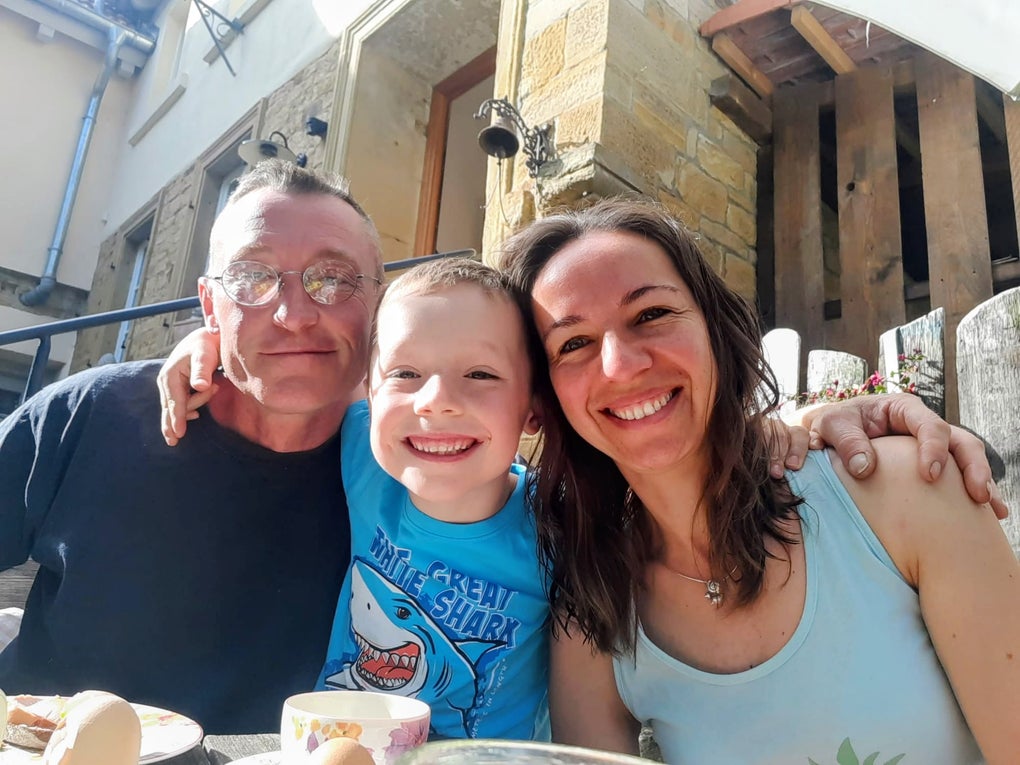 Patient Micha mit seiner Familie, links der Sohn und rechts die Frau. Alle sitzen draußen vor einem Tisch und lächeln Patient Micha mit seiner Familie, links der Sohn und rechts die Frau. Alle sitzen draußen vor einem Tisch und lächeln