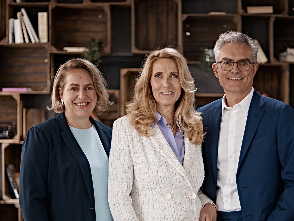Porträt von Dr. Elke Neujahr, Dr. Deborah Buk und Stephan Schumacher im Business-Outfit vor einem Regal mit Büchern und Dekoration. Porträt von Dr. Elke Neujahr, Dr. Deborah Buk und Stephan Schumacher im Business-Outfit vor einem Regal mit Büchern und Dekoration.