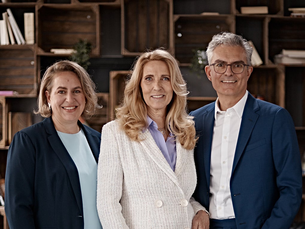 Porträt von Dr. Elke Neujahr, Dr. Deborah Buk und Stephan Schumacher im Business-Outfit vor einem Regal mit Büchern und Dekoration. Porträt von Dr. Elke Neujahr, Dr. Deborah Buk und Stephan Schumacher im Business-Outfit vor einem Regal mit Büchern und Dekoration.