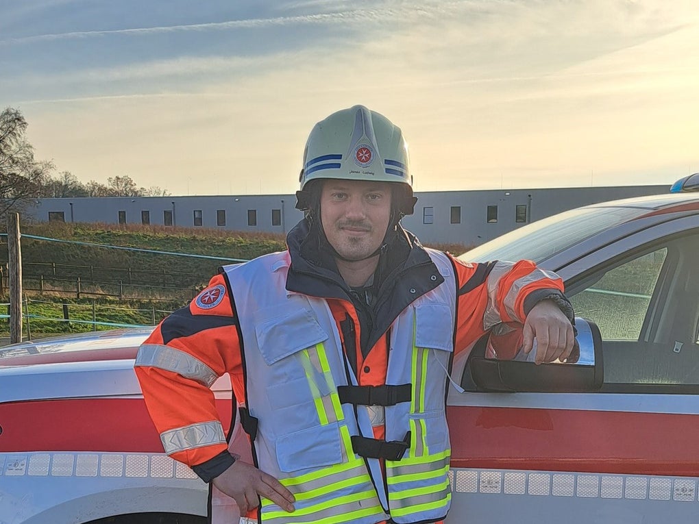 Eine Person in leuchtend orangefarbener Einsatzkleidung mit reflektierenden Streifen steht neben einem Einsatzfahrzeug der Johanniter. Sie trägt einen Schutzhelm mit Johanniter-Logo und lehnt mit dem rechten Arm an der geöffneten Fahrertür. Im Hintergrund sind ein freies Feld und ein langgezogenes Gebäude zu sehen, der Himmel ist teils bewölkt. Eine Person in leuchtend orangefarbener Einsatzkleidung mit reflektierenden Streifen steht neben einem Einsatzfahrzeug der Johanniter. Sie trägt einen Schutzhelm mit Johanniter-Logo und lehnt mit dem rechten Arm an der geöffneten Fahrertür. Im Hintergrund sind ein freies Feld und ein langgezogenes Gebäude zu sehen, der Himmel ist teils bewölkt.