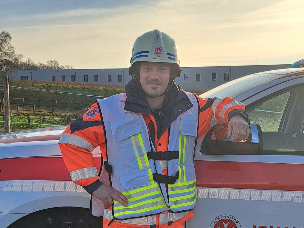 Eine Person in leuchtend orangefarbener Einsatzkleidung mit reflektierenden Streifen steht neben einem Einsatzfahrzeug der Johanniter. Sie trägt einen Schutzhelm mit Johanniter-Logo und lehnt mit dem rechten Arm an der geöffneten Fahrertür. Im Hintergrund sind ein freies Feld und ein langgezogenes Gebäude zu sehen, der Himmel ist teils bewölkt. Eine Person in leuchtend orangefarbener Einsatzkleidung mit reflektierenden Streifen steht neben einem Einsatzfahrzeug der Johanniter. Sie trägt einen Schutzhelm mit Johanniter-Logo und lehnt mit dem rechten Arm an der geöffneten Fahrertür. Im Hintergrund sind ein freies Feld und ein langgezogenes Gebäude zu sehen, der Himmel ist teils bewölkt.