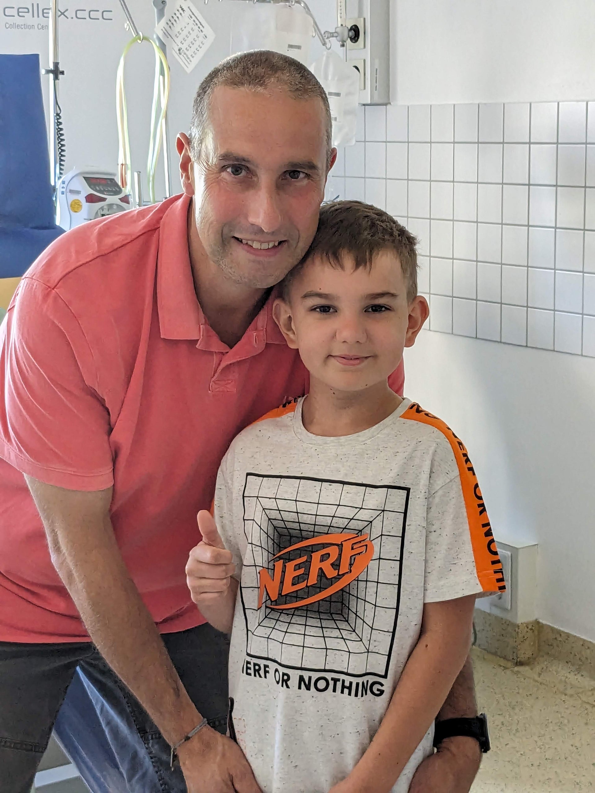 Ein Mann in einem rosa Poloshirt und ein Junge in einem weißen T-Shirt mit „NERF“-Aufdruck stehen nebeneinander in einem Klinikzimmer. Beide lächeln in die Kamera, der Junge zeigt den Daumen hoch. Im Hintergrund sind medizinische Geräte, eine Fliese an der Wand und ein Infusionsständer zu sehen. Ein Mann in einem rosa Poloshirt und ein Junge in einem weißen T-Shirt mit „NERF“-Aufdruck stehen nebeneinander in einem Klinikzimmer. Beide lächeln in die Kamera, der Junge zeigt den Daumen hoch. Im Hintergrund sind medizinische Geräte, eine Fliese an der Wand und ein Infusionsständer zu sehen.