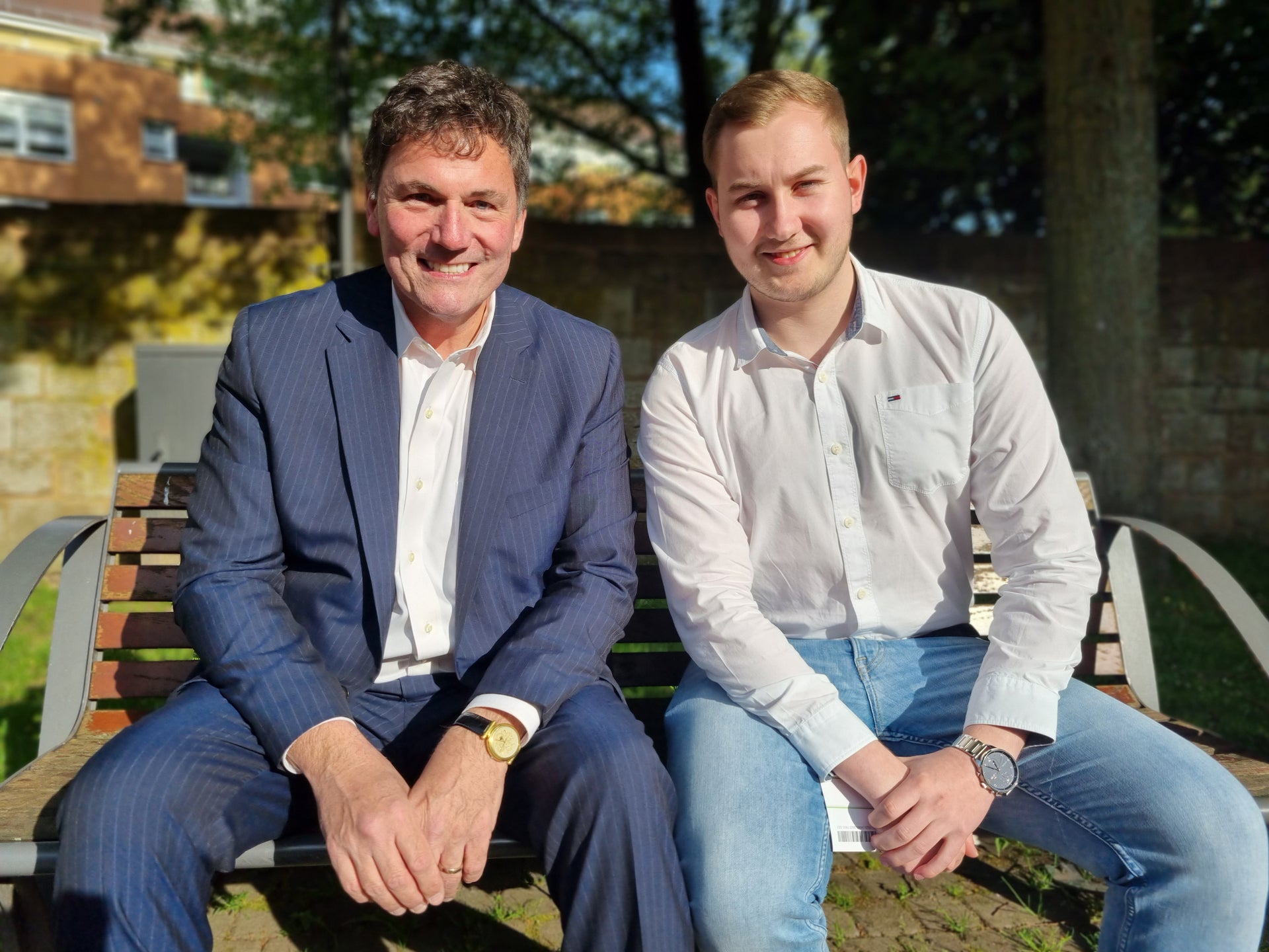 Zwei Männer sitzen nebeneinander auf einer Holzbank im Freien und blicken in die Kamera. Der Mann links trägt einen dunkelblauen Anzug, der Mann rechts ein weißes Hemd und eine blaue Jeans. Beide haben die Hände gefaltet oder auf dem Schoß. Im Hintergrund sind Bäume, eine Mauer und ein Gebäude zu sehen. Die Sonne scheint. Zwei Männer sitzen nebeneinander auf einer Holzbank im Freien und blicken in die Kamera. Der Mann links trägt einen dunkelblauen Anzug, der Mann rechts ein weißes Hemd und eine blaue Jeans. Beide haben die Hände gefaltet oder auf dem Schoß. Im Hintergrund sind Bäume, eine Mauer und ein Gebäude zu sehen. Die Sonne scheint.