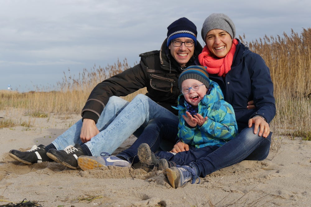 Auf dem Bild sieht man ein gemeinsames Bild von Nils mit seinen Eltern. Sie sitzen lachend auf einem Sandboden. 