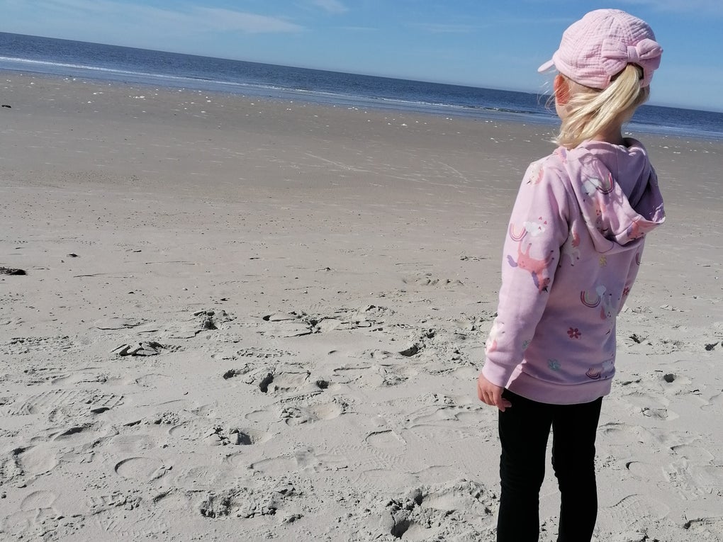 Ein blondes Mädchen mit rosa Cap und Hoodie steht mit dem Rücken zur Kamera und schaut aufs Meer. Ein blondes Mädchen mit rosa Cap und Hoodie steht mit dem Rücken zur Kamera und schaut aufs Meer.