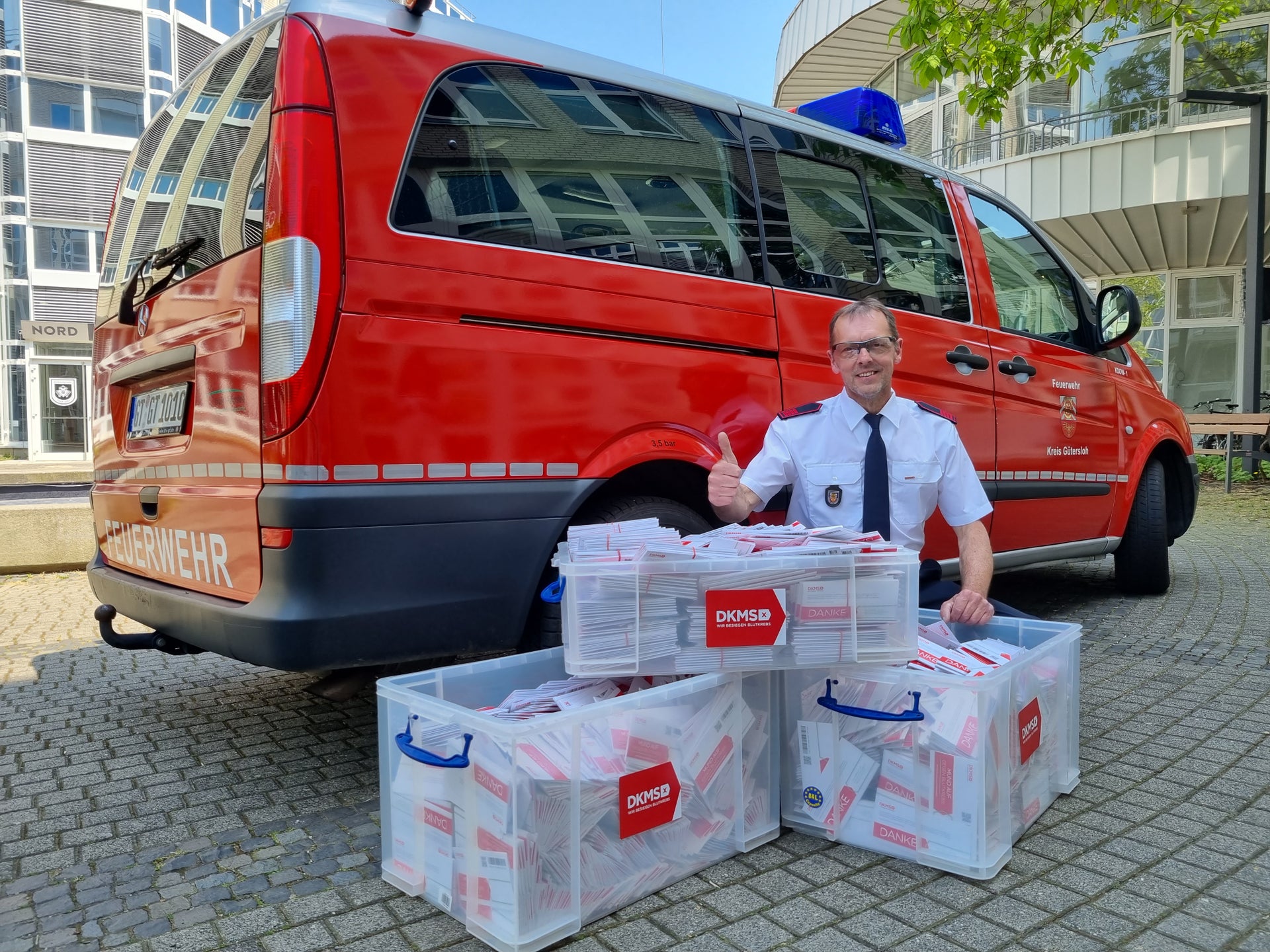Ein Mann in weißem Diensthemd und schwarzer Krawatte sitzt auf dem Boden vor einem roten Feuerwehrfahrzeug. Er zeigt den Daumen nach oben. Vor ihm stehen drei transparente Kunststoffboxen, die mit DKMS-Registrierungssets gefüllt sind. Im Hintergrund sind ein modernes Gebäude mit Glasfassade und ein Baum zu sehen.
Ein Mann in weißem Diensthemd und schwarzer Krawatte sitzt auf dem Boden vor einem roten Feuerwehrfahrzeug. Er zeigt den Daumen nach oben. Vor ihm stehen drei transparente Kunststoffboxen, die mit DKMS-Registrierungssets gefüllt sind. Im Hintergrund sind ein modernes Gebäude mit Glasfassade und ein Baum zu sehen.