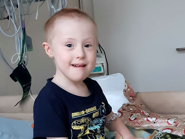 Junge mit kurzem Haar sitzt in einem Krankenhausbett und lächelt. Er trägt ein dunkles T-Shirt mit Dinosaurier-Aufdruck. Im Hintergrund sind medizinische Geräte und Schläuche zu sehen. Neben ihm liegt Geschenkpapier mit Weihnachtsmotiven. Junge mit kurzem Haar sitzt in einem Krankenhausbett und lächelt. Er trägt ein dunkles T-Shirt mit Dinosaurier-Aufdruck. Im Hintergrund sind medizinische Geräte und Schläuche zu sehen. Neben ihm liegt Geschenkpapier mit Weihnachtsmotiven.