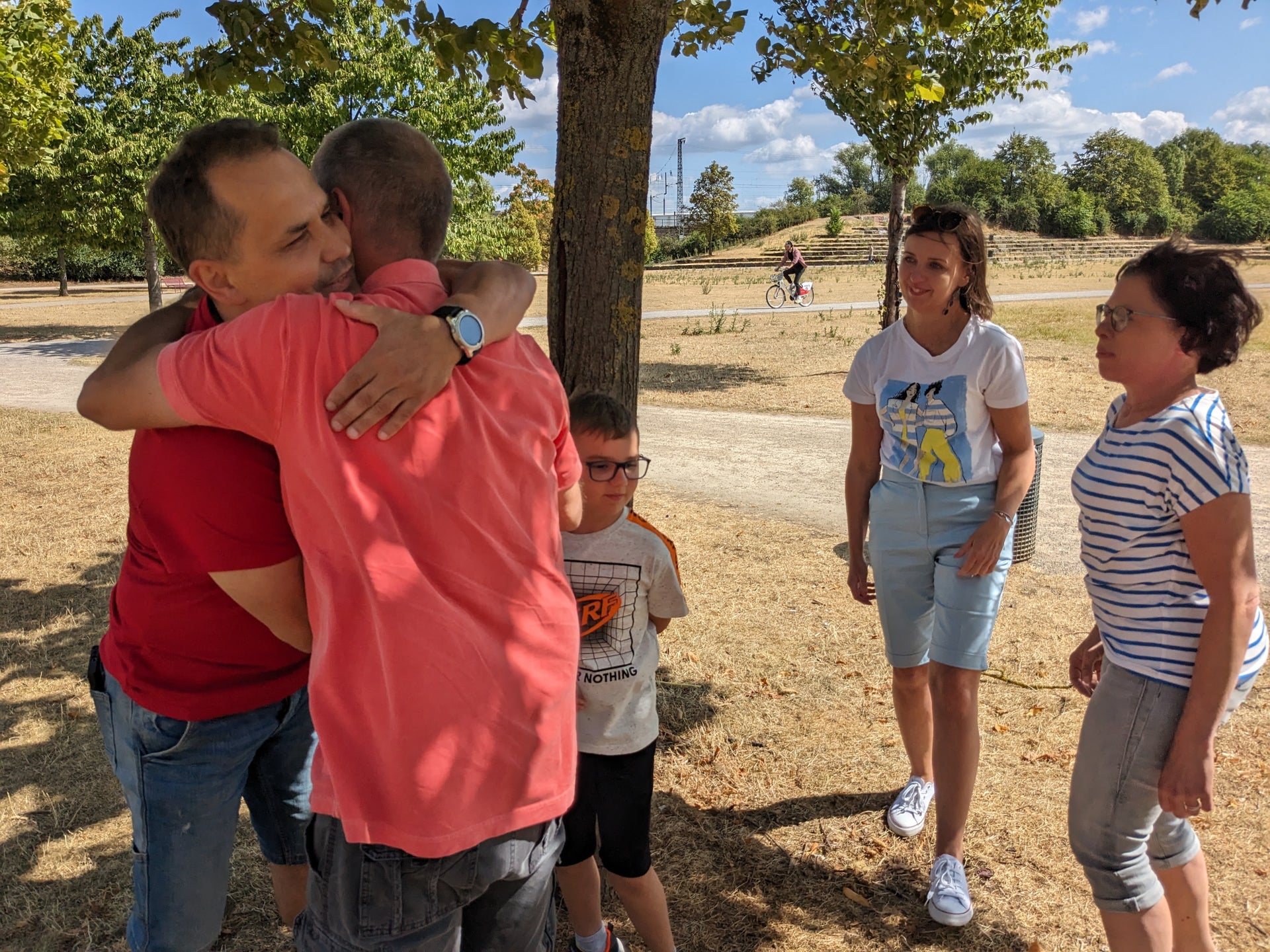 Zwei Männer umarmen sich unter einem Baum in einem Park. Einer trägt ein rotes, der andere ein rosafarbenes T-Shirt. Neben ihnen stehen ein Junge mit Brille, eine Frau in Latzhose und T-Shirt sowie eine Frau in gestreiftem Shirt. Der Boden ist trocken und mit hellem Gras bedeckt, im Hintergrund sind ein Weg, Bäume und weitere Personen sichtbar. Zwei Männer umarmen sich unter einem Baum in einem Park. Einer trägt ein rotes, der andere ein rosafarbenes T-Shirt. Neben ihnen stehen ein Junge mit Brille, eine Frau in Latzhose und T-Shirt sowie eine Frau in gestreiftem Shirt. Der Boden ist trocken und mit hellem Gras bedeckt, im Hintergrund sind ein Weg, Bäume und weitere Personen sichtbar.