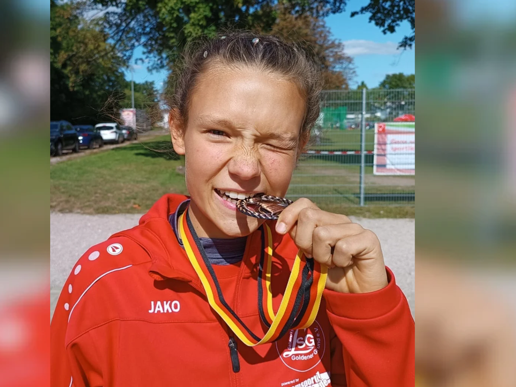 Ein Mädchen trägt eine rote Trainingsjacke und beißt auf eine Bronzemedaille. Ein Mädchen trägt eine rote Trainingsjacke und beißt auf eine Bronzemedaille.