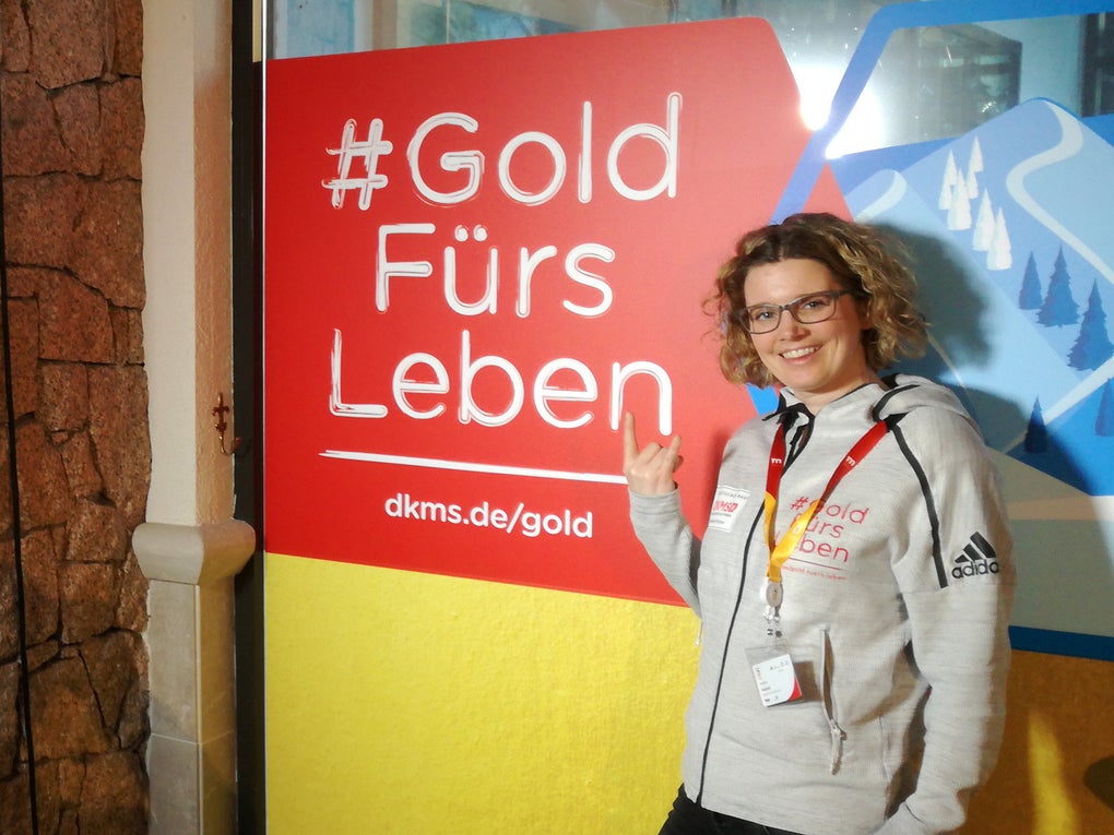Eine Frau steht vor einem Schild der DKMS mit dem Text #Gold Fürs Leben Eine Frau steht vor einem Schild der DKMS mit dem Text #Gold Fürs Leben