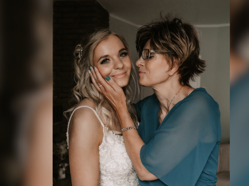 Eine ältere Frau trägt ein grünblaues Kleid und hält das Gesicht einer jungen, ein Hochzeitskleid tragenden Frau in den Händen. Eine ältere Frau trägt ein grünblaues Kleid und hält das Gesicht einer jungen, ein Hochzeitskleid tragenden Frau in den Händen.