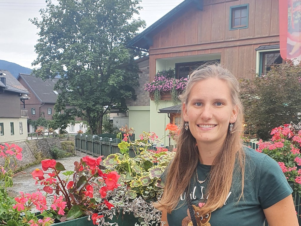 Eine junge Frau steht lächelnd an einem grünen Geländer, das mit pinken und roten Blumen geschmückt ist. Im Hintergrund sind ein kleiner Fluss, ein Haus mit Blumenkästen am Balkon und ein Baum zu sehen. Die Frau trägt ein dunkelgrünes T-Shirt, eine schwarze Hose und eine schwarze Umhängetasche. Es ist bewölkt. Eine junge Frau steht lächelnd an einem grünen Geländer, das mit pinken und roten Blumen geschmückt ist. Im Hintergrund sind ein kleiner Fluss, ein Haus mit Blumenkästen am Balkon und ein Baum zu sehen. Die Frau trägt ein dunkelgrünes T-Shirt, eine schwarze Hose und eine schwarze Umhängetasche. Es ist bewölkt.