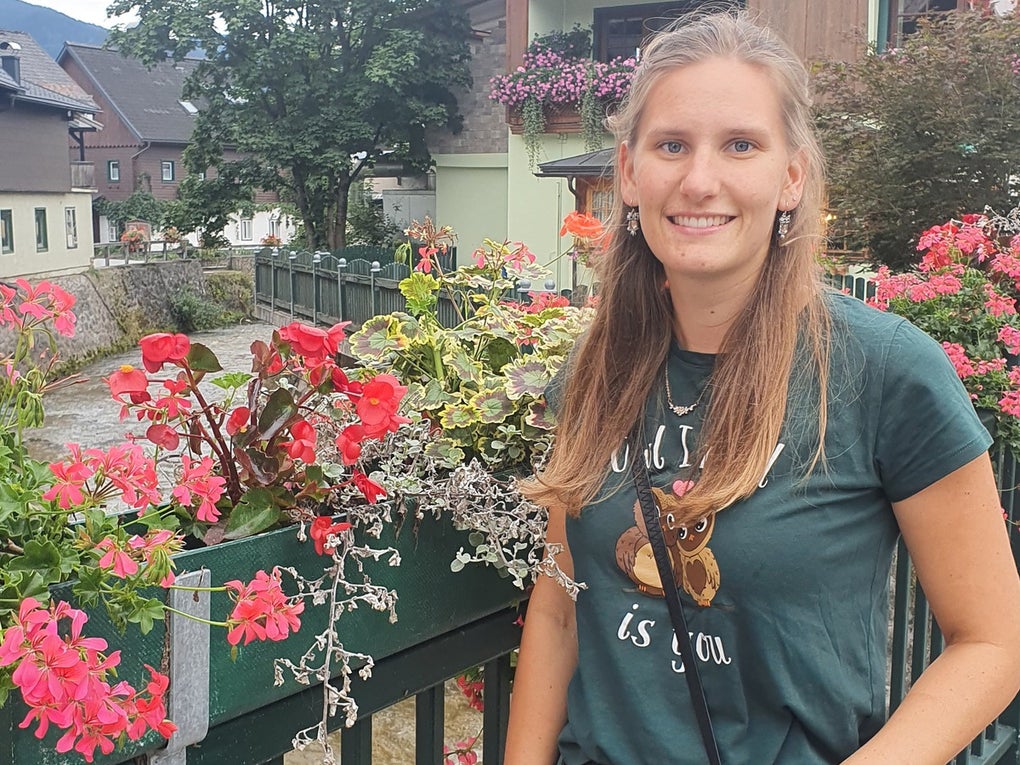 Eine junge Frau steht lächelnd an einem grünen Geländer, das mit pinken und roten Blumen geschmückt ist. Im Hintergrund sind ein kleiner Fluss, ein Haus mit Blumenkästen am Balkon und ein Baum zu sehen. Die Frau trägt ein dunkelgrünes T-Shirt, eine schwarze Hose und eine schwarze Umhängetasche. Es ist bewölkt. Eine junge Frau steht lächelnd an einem grünen Geländer, das mit pinken und roten Blumen geschmückt ist. Im Hintergrund sind ein kleiner Fluss, ein Haus mit Blumenkästen am Balkon und ein Baum zu sehen. Die Frau trägt ein dunkelgrünes T-Shirt, eine schwarze Hose und eine schwarze Umhängetasche. Es ist bewölkt.