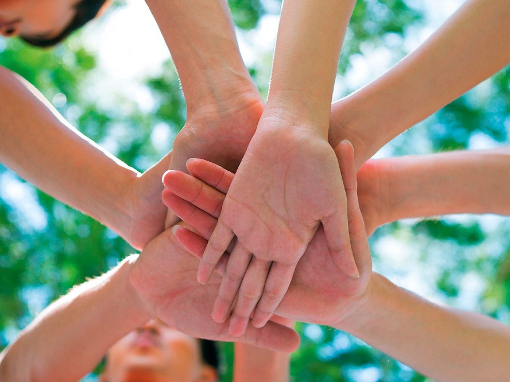 Mehrere Menschen legen im Kreis stehend ihre Hände übereinander, als Zeichen von Teamgeist und Zusammenhalt. Der Hintergrund zeigt grünes Laub und blauen Himmel, die Perspektive ist von unten aufgenommen.  Mehrere Menschen legen im Kreis stehend ihre Hände übereinander, als Zeichen von Teamgeist und Zusammenhalt. Der Hintergrund zeigt grünes Laub und blauen Himmel, die Perspektive ist von unten aufgenommen.