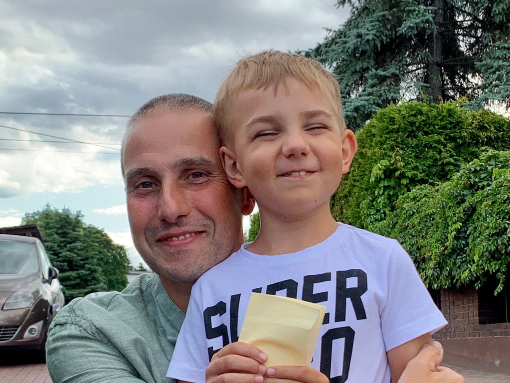Ein Mann hält ein Kind im Arm, beide lächeln in die Kamera. Der Junge trägt ein weißes T-Shirt mit schwarzer Aufschrift und hält einen gelben Umschlag in den Händen. Der Mann trägt ein grünes T-Shirt. Im Hintergrund sind ein gepflasterter Hof, ein Auto, grüne Sträucher und Bäume sowie ein bewölkter Himmel zu sehen. Ein Mann hält ein Kind im Arm, beide lächeln in die Kamera. Der Junge trägt ein weißes T-Shirt mit schwarzer Aufschrift und hält einen gelben Umschlag in den Händen. Der Mann trägt ein grünes T-Shirt. Im Hintergrund sind ein gepflasterter Hof, ein Auto, grüne Sträucher und Bäume sowie ein bewölkter Himmel zu sehen.