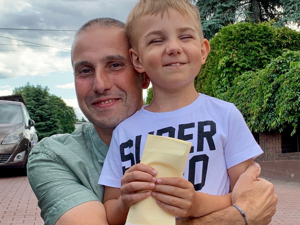 Ein Mann hält ein Kind im Arm, beide lächeln in die Kamera. Der Junge trägt ein weißes T-Shirt mit schwarzer Aufschrift und hält einen gelben Umschlag in den Händen. Der Mann trägt ein grünes T-Shirt. Im Hintergrund sind ein gepflasterter Hof, ein Auto, grüne Sträucher und Bäume sowie ein bewölkter Himmel zu sehen. Ein Mann hält ein Kind im Arm, beide lächeln in die Kamera. Der Junge trägt ein weißes T-Shirt mit schwarzer Aufschrift und hält einen gelben Umschlag in den Händen. Der Mann trägt ein grünes T-Shirt. Im Hintergrund sind ein gepflasterter Hof, ein Auto, grüne Sträucher und Bäume sowie ein bewölkter Himmel zu sehen.