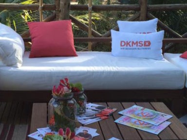 Eine weiße Couch mit Kissen, auf zwei Kissen sind DKMS Logos gedrückt. Davor ein Tisch mit Blumen und Zeitschriften Eine weiße Couch mit Kissen, auf zwei Kissen sind DKMS Logos gedrückt. Davor ein Tisch mit Blumen und Zeitschriften