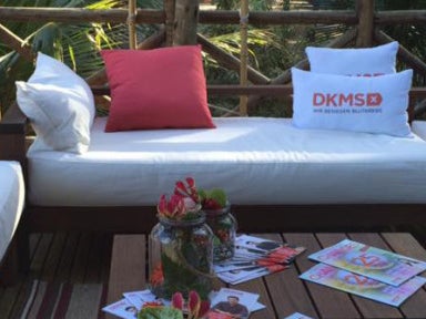 Eine weiße Couch mit Kissen, auf zwei Kissen sind DKMS Logos gedrückt. Davor ein Tisch mit Blumen und Zeitschriften Eine weiße Couch mit Kissen, auf zwei Kissen sind DKMS Logos gedrückt. Davor ein Tisch mit Blumen und Zeitschriften