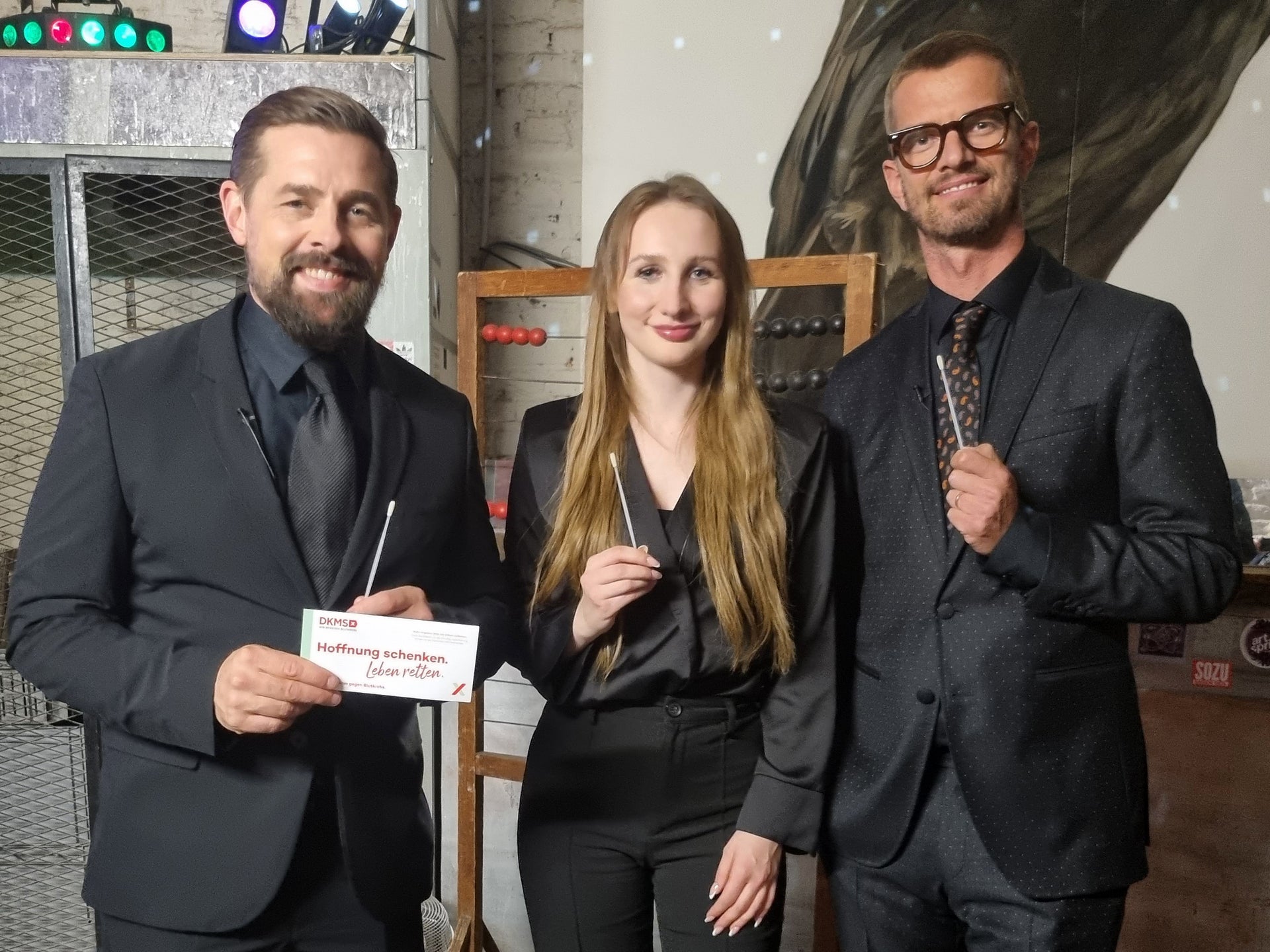 Zwei Männer und eine Frau stehen nebeneinander und blicken lächelnd in die Kamera. Alle drei tragen dunkle Kleidung. Die Frau und der Mann rechts halten jeweils ein Wattestäbchen in der Hand. Der Mann links hält zusätzlich ein DKMS-Kärtchen mit der Aufschrift „Mund auf. Stäbchen rein. Spender sein.“. Im Hintergrund sind bunte Lichter und metallische Regale zu sehen. Zwei Männer und eine Frau stehen nebeneinander und blicken lächelnd in die Kamera. Alle drei tragen dunkle Kleidung. Die Frau und der Mann rechts halten jeweils ein Wattestäbchen in der Hand. Der Mann links hält zusätzlich ein DKMS-Kärtchen mit der Aufschrift „Mund auf. Stäbchen rein. Spender sein.“. Im Hintergrund sind bunte Lichter und metallische Regale zu sehen.