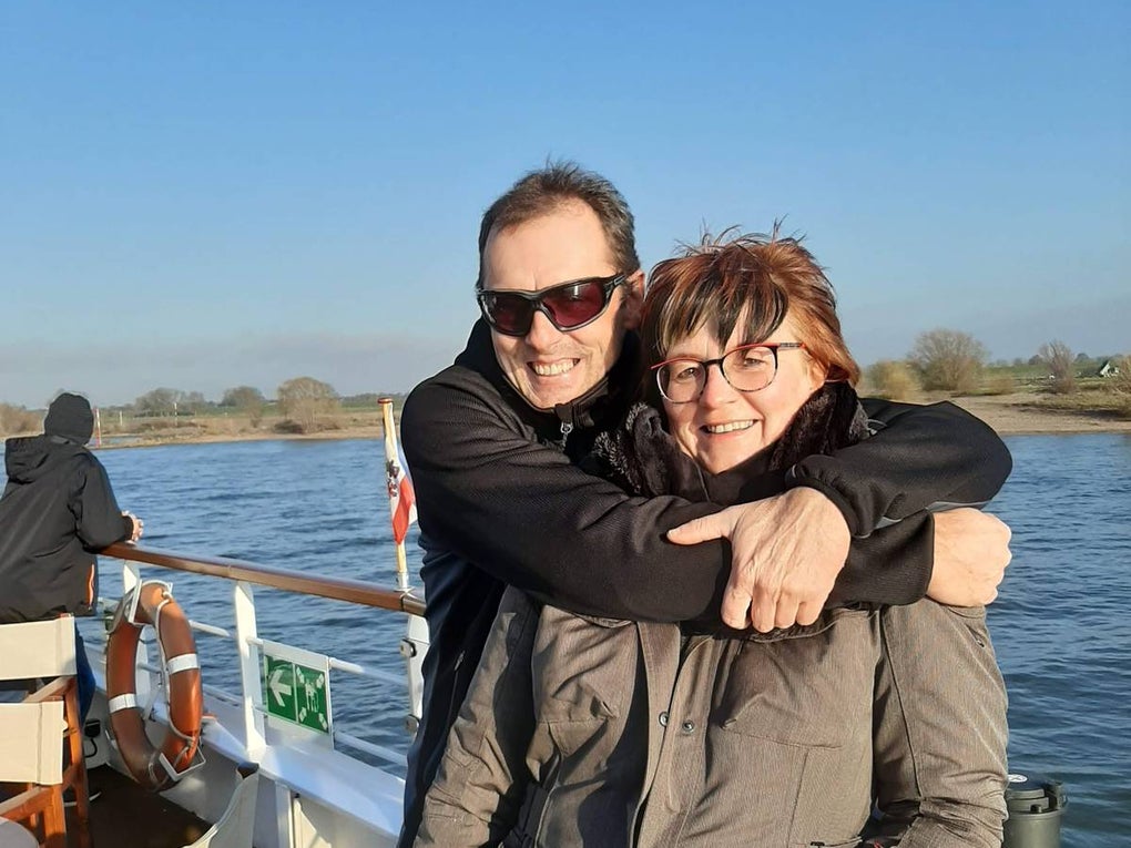 Ein Mann steht auf einem Schiff hinter einer Frau und umarmt sie von hinten. Beide tragen Jacken, der Mann eine Sonnenbrille. Sie stehen an der Reling, im Hintergrund ist Wasser, Ufer mit Bäumen und blauer Himmel zu sehen. Auf dem Deck stehen mehrere Stühle. Ein Mann steht auf einem Schiff hinter einer Frau und umarmt sie von hinten. Beide tragen Jacken, der Mann eine Sonnenbrille. Sie stehen an der Reling, im Hintergrund ist Wasser, Ufer mit Bäumen und blauer Himmel zu sehen. Auf dem Deck stehen mehrere Stühle.