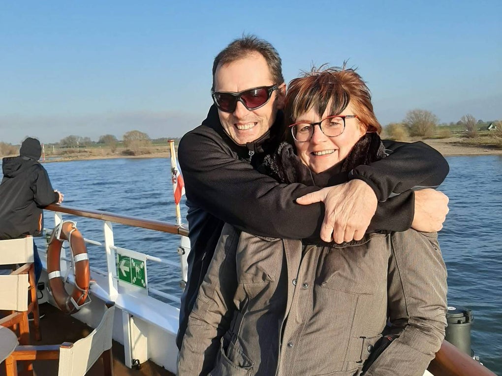 Ein Mann steht auf einem Schiff hinter einer Frau und umarmt sie von hinten. Beide tragen Jacken, der Mann eine Sonnenbrille. Sie stehen an der Reling, im Hintergrund ist Wasser, Ufer mit Bäumen und blauer Himmel zu sehen. Auf dem Deck stehen mehrere Stühle. Ein Mann steht auf einem Schiff hinter einer Frau und umarmt sie von hinten. Beide tragen Jacken, der Mann eine Sonnenbrille. Sie stehen an der Reling, im Hintergrund ist Wasser, Ufer mit Bäumen und blauer Himmel zu sehen. Auf dem Deck stehen mehrere Stühle.