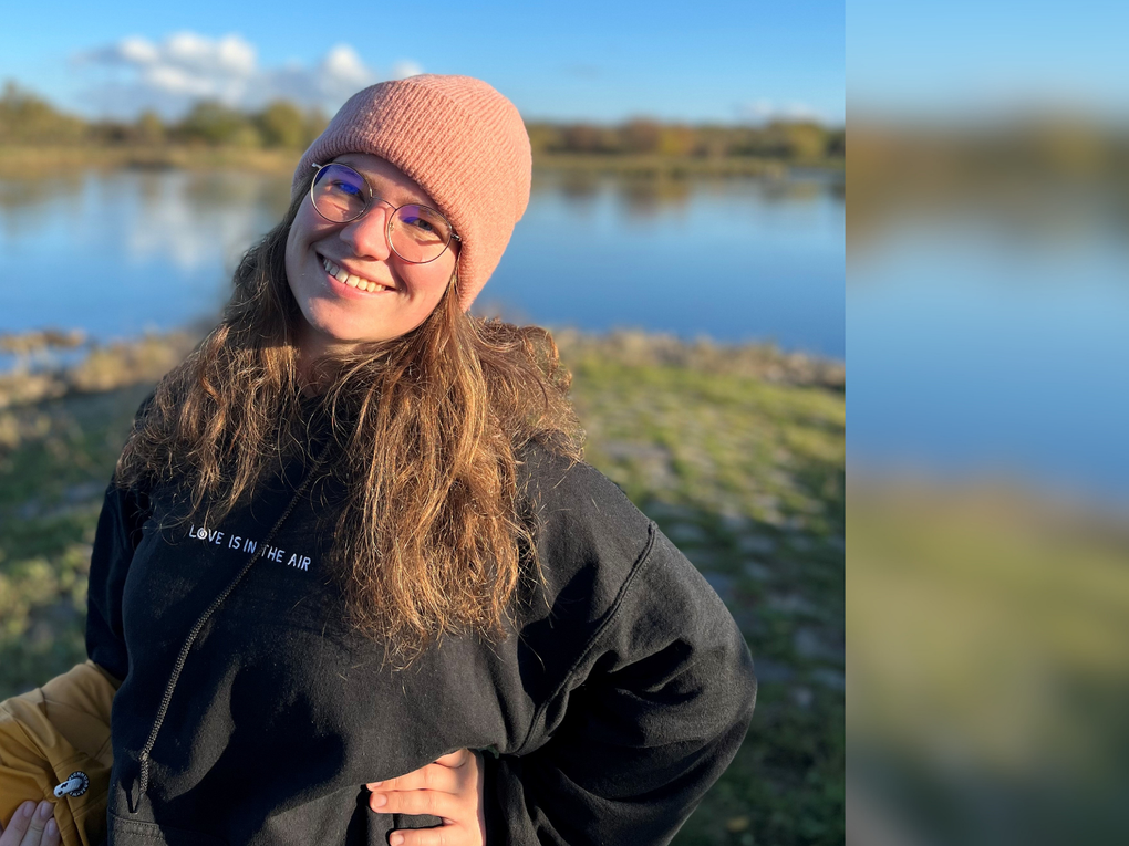 Eine Frau trägt eine rosafarbene Mütze und einen schwarzen Kapuzenpullover. Sie steht am Ufer eines Gewässers. Eine Frau trägt eine rosafarbene Mütze und einen schwarzen Kapuzenpullover. Sie steht am Ufer eines Gewässers.