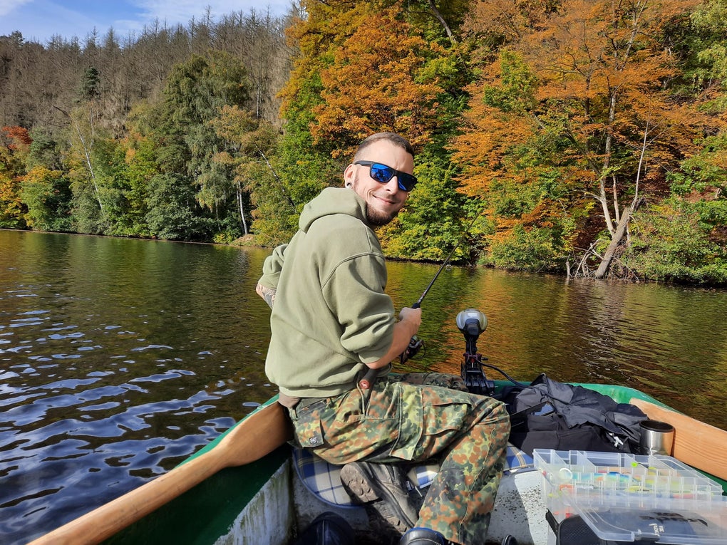 ein Mann sitzt auf einem Ruderboot mitten auf einem See. Im Hintergrund sieht man herbstliche Laubbäume ein Mann sitzt auf einem Ruderboot mitten auf einem See. Im Hintergrund sieht man herbstliche Laubbäume