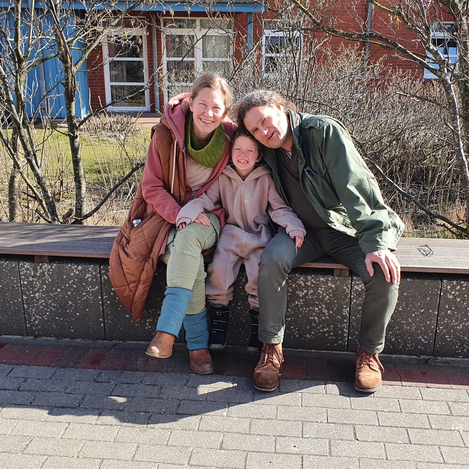 Ein fröhliches Familienfoto im Freien. Zwei Erwachsene und ein Kind sitzen eng aneinandergeschmiegt auf einer Holzbank und lächeln in die Kamera. Die Erwachsene links trägt eine orangefarbene Jacke und einen grünen Schal, das Kind in der Mitte trägt einen beigefarbenen Einteiler, der Erwachsene rechts eine grüne Jacke. Im Hintergrund sind kahle Büsche, ein rotes Backsteingebäude und Fenster mit weißen Rahmen zu sehen. Ein fröhliches Familienfoto im Freien. Zwei Erwachsene und ein Kind sitzen eng aneinandergeschmiegt auf einer Holzbank und lächeln in die Kamera. Die Erwachsene links trägt eine orangefarbene Jacke und einen grünen Schal, das Kind in der Mitte trägt einen beigefarbenen Einteiler, der Erwachsene rechts eine grüne Jacke. Im Hintergrund sind kahle Büsche, ein rotes Backsteingebäude und Fenster mit weißen Rahmen zu sehen.