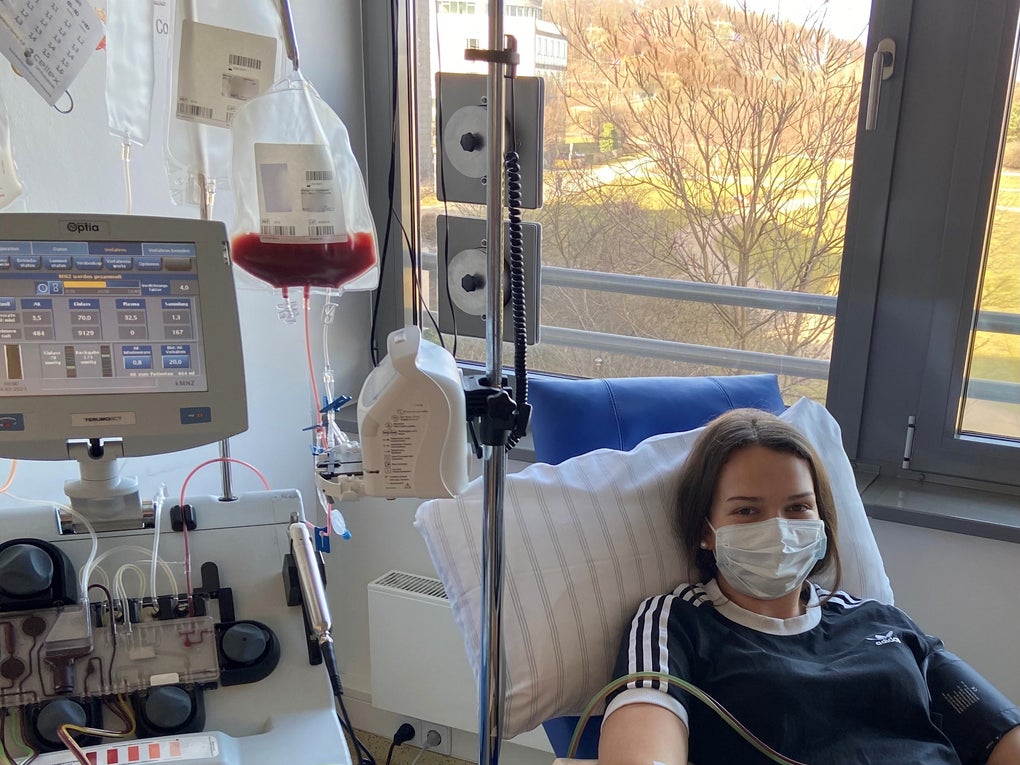 Eine Frau liegt in einem medizinischen Behandlungsstuhl und spendet periphere Blutstammzellen. Sie trägt eine medizinische Maske und ein dunkles T-Shirt. Ihr rechter Arm ist an eine Apheresemaschine angeschlossen. Neben ihr stehen Infusionsbeutel, ein Monitor und medizinisches Zubehör. Durch das Fenster ist eine Landschaft mit Bäumen und Wiese zu sehen. Eine Frau liegt in einem medizinischen Behandlungsstuhl und spendet periphere Blutstammzellen. Sie trägt eine medizinische Maske und ein dunkles T-Shirt. Ihr rechter Arm ist an eine Apheresemaschine angeschlossen. Neben ihr stehen Infusionsbeutel, ein Monitor und medizinisches Zubehör. Durch das Fenster ist eine Landschaft mit Bäumen und Wiese zu sehen.