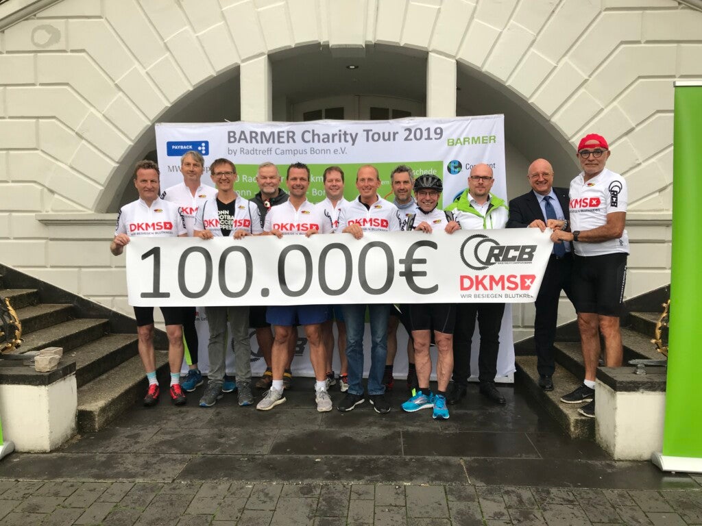 Eine Gruppe von Menschen, die Trikots mit DKMS Logo trägt. Sie tragen ein Banner mit dem Aufdruck 100.000 € Eine Gruppe von Menschen, die Trikots mit DKMS Logo trägt. Sie tragen ein Banner mit dem Aufdruck 100.000 €