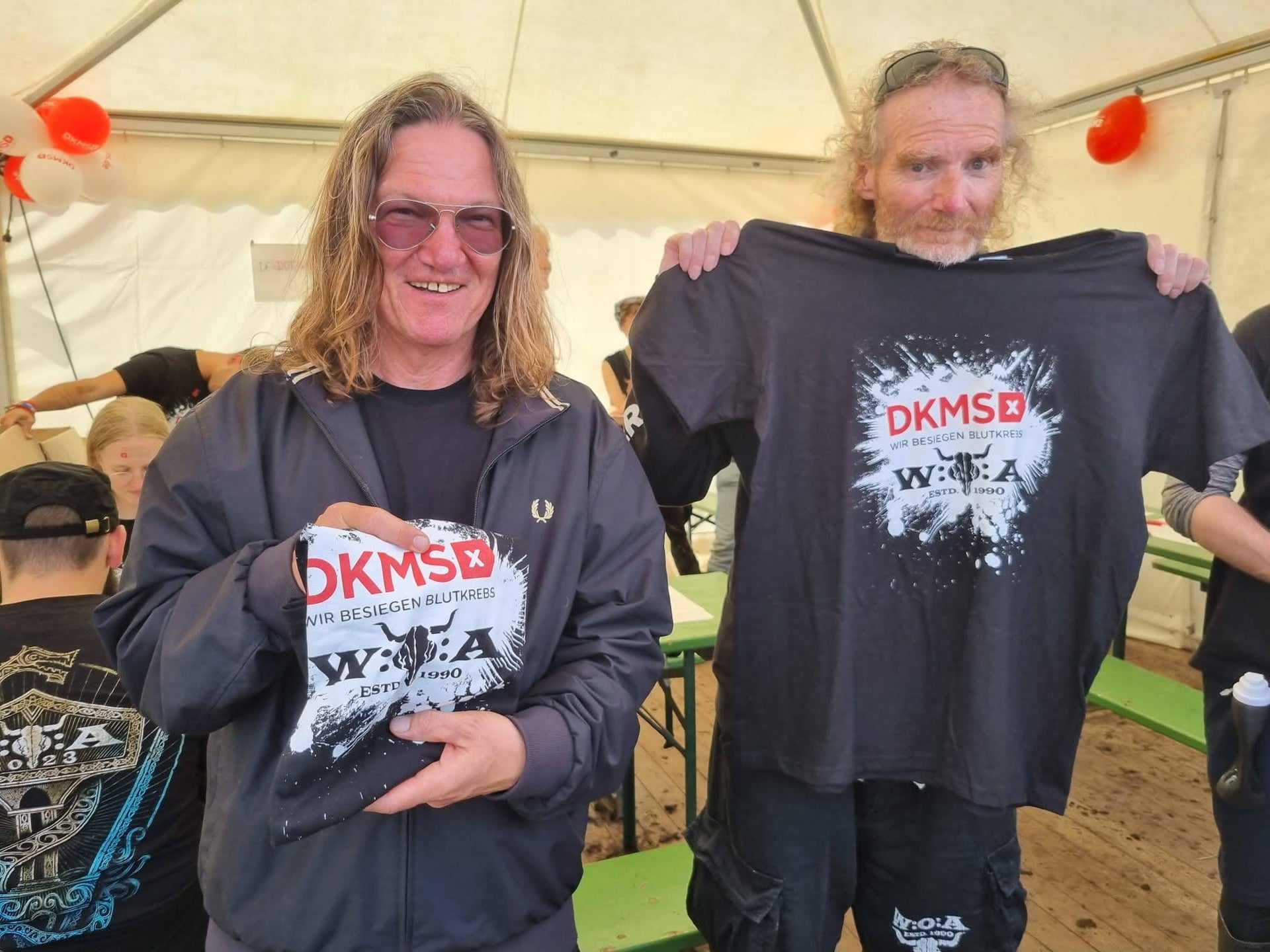 Zwei Männer stehen in einem Zelt und halten jeweils ein schwarzes DKMS-T-Shirt mit Wacken-Design hoch. Beide tragen dunkle Kleidung, einer hat lange glatte Haare und Sonnenbrille, der andere lockiges, schulterlanges Haar. Im Hintergrund sitzen mehrere Personen an Bierzeltgarnituren, über ihnen hängen rote und weiße Luftballons. Zwei Männer stehen in einem Zelt und halten jeweils ein schwarzes DKMS-T-Shirt mit Wacken-Design hoch. Beide tragen dunkle Kleidung, einer hat lange glatte Haare und Sonnenbrille, der andere lockiges, schulterlanges Haar. Im Hintergrund sitzen mehrere Personen an Bierzeltgarnituren, über ihnen hängen rote und weiße Luftballons.