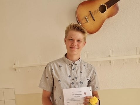 Ein Jugendlicher steht in einem Raum vor einer hellen Wand mit Hakenleiste und hält ein Dokument sowie eine gelbe Rose in der Hand. Er trägt ein kurzärmliges Hemd mit Palmenmuster und eine kurze Jeanshose. Über ihm hängt eine Akustikgitarre an der Wand. Ein Jugendlicher steht in einem Raum vor einer hellen Wand mit Hakenleiste und hält ein Dokument sowie eine gelbe Rose in der Hand. Er trägt ein kurzärmliges Hemd mit Palmenmuster und eine kurze Jeanshose. Über ihm hängt eine Akustikgitarre an der Wand.