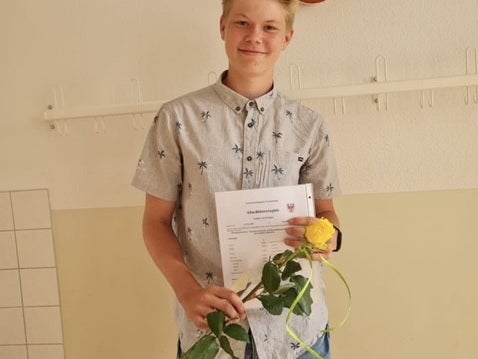 Ein Jugendlicher steht in einem Raum vor einer hellen Wand mit Hakenleiste und hält ein Dokument sowie eine gelbe Rose in der Hand. Er trägt ein kurzärmliges Hemd mit Palmenmuster und eine kurze Jeanshose. Über ihm hängt eine Akustikgitarre an der Wand. Ein Jugendlicher steht in einem Raum vor einer hellen Wand mit Hakenleiste und hält ein Dokument sowie eine gelbe Rose in der Hand. Er trägt ein kurzärmliges Hemd mit Palmenmuster und eine kurze Jeanshose. Über ihm hängt eine Akustikgitarre an der Wand.