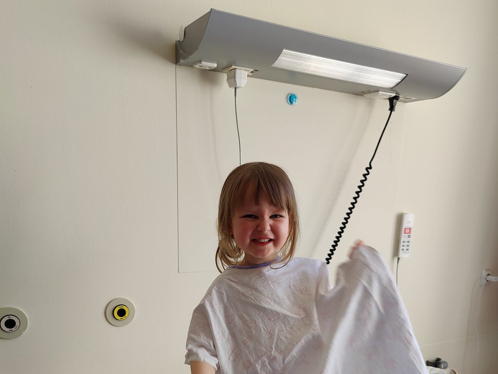 Ein kleines Kind steht lachend in einem Krankenhausbett und hält ein Tuch oder eine Decke in der Hand. Auf dem Bett liegen Kuscheltiere und Kissen. Ein kleines Kind steht lachend in einem Krankenhausbett und hält ein Tuch oder eine Decke in der Hand. Auf dem Bett liegen Kuscheltiere und Kissen.