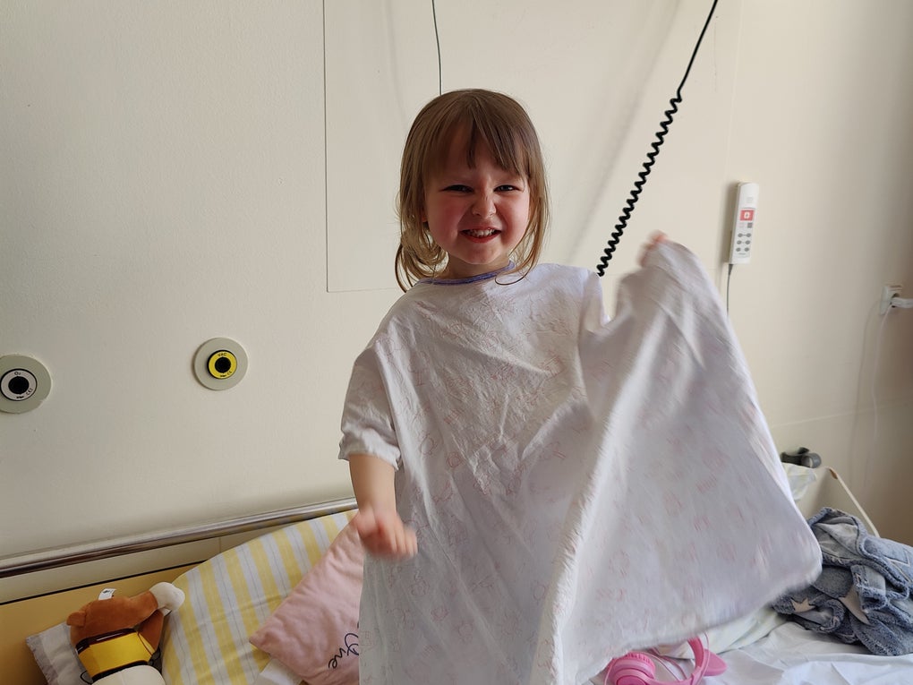 Ein kleines Kind steht lachend in einem Krankenhausbett und hält ein Tuch oder eine Decke in der Hand. Auf dem Bett liegen Kuscheltiere und Kissen. Ein kleines Kind steht lachend in einem Krankenhausbett und hält ein Tuch oder eine Decke in der Hand. Auf dem Bett liegen Kuscheltiere und Kissen.