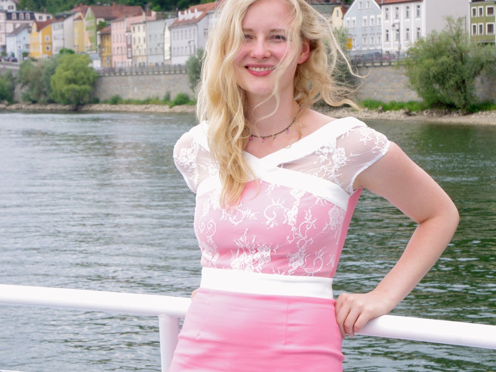 Eine Frau in einem rosa Kleid lehnt an einem Geländer auf einem Schiff, im Hintergrund ist Wasser Eine Frau in einem rosa Kleid lehnt an einem Geländer auf einem Schiff, im Hintergrund ist Wasser