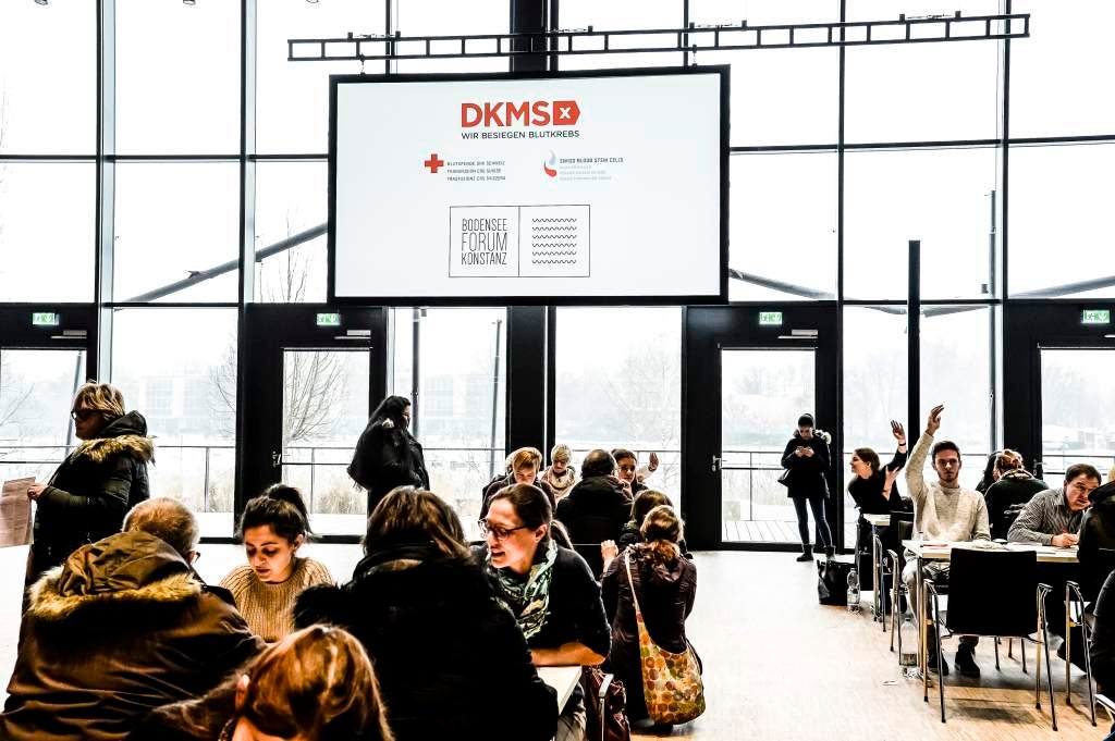 Eine Gruppe von Menschen, die in einem Raum mit einem großen Bildschirm sitzen. Auf dem Bildschirm ist ein DKMS Logo Eine Gruppe von Menschen, die in einem Raum mit einem großen Bildschirm sitzen. Auf dem Bildschirm ist ein DKMS Logo