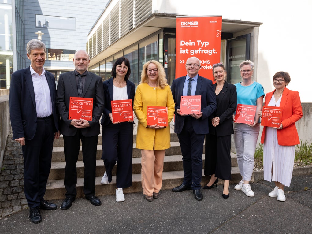 Acht Personen stehen nebeneinander vor einem Gebäude. Sechs von ihnen halten rote DKMS-Schilder mit der Aufschrift „Wir retten Leben“. Im Hintergrund hängt ein großes rotes DKMS-Banner mit dem Text „Dein Typ ist gefragt“. Alle Personen blicken in die Kamera und lächeln. Acht Personen stehen nebeneinander vor einem Gebäude. Sechs von ihnen halten rote DKMS-Schilder mit der Aufschrift „Wir retten Leben“. Im Hintergrund hängt ein großes rotes DKMS-Banner mit dem Text „Dein Typ ist gefragt“. Alle Personen blicken in die Kamera und lächeln.