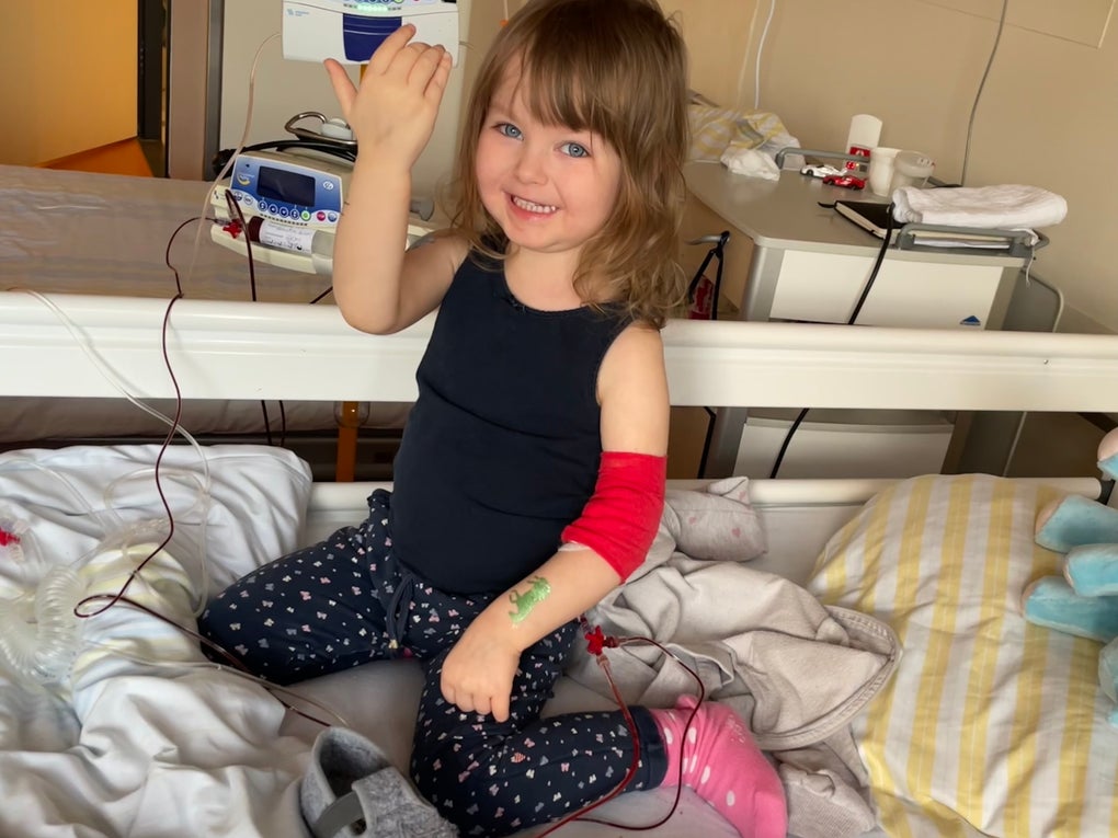 Ein kleines Mädchen sitzt auf einem Krankenhausbett und lächelt in die Kamera. Sie trägt ein dunkles Oberteil, gepunktete Leggings, eine pinke Socke und hat einen roten Verband am rechten Arm. An ihrem Arm ist ein Zugang gelegt, der mit einer Infusionsleitung verbunden ist. Im Hintergrund sind medizinisches Gerät, ein Nachttisch und weitere Gegenstände zu sehen.
Ein kleines Mädchen sitzt auf einem Krankenhausbett und lächelt in die Kamera. Sie trägt ein dunkles Oberteil, gepunktete Leggings, eine pinke Socke und hat einen roten Verband am rechten Arm. An ihrem Arm ist ein Zugang gelegt, der mit einer Infusionsleitung verbunden ist. Im Hintergrund sind medizinisches Gerät, ein Nachttisch und weitere Gegenstände zu sehen.