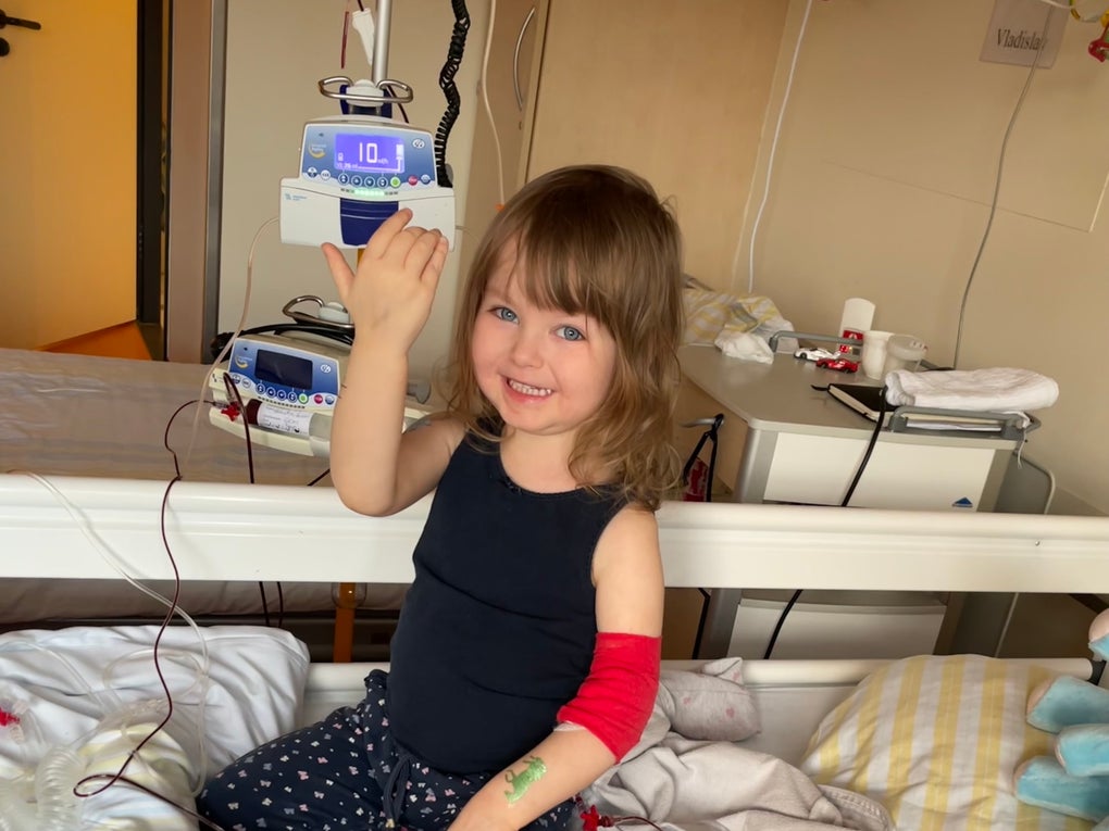 Ein kleines Mädchen sitzt auf einem Krankenhausbett und lächelt in die Kamera. Sie trägt ein dunkles Oberteil, gepunktete Leggings, eine pinke Socke und hat einen roten Verband am rechten Arm. An ihrem Arm ist ein Zugang gelegt, der mit einer Infusionsleitung verbunden ist. Im Hintergrund sind medizinisches Gerät, ein Nachttisch und weitere Gegenstände zu sehen.
Ein kleines Mädchen sitzt auf einem Krankenhausbett und lächelt in die Kamera. Sie trägt ein dunkles Oberteil, gepunktete Leggings, eine pinke Socke und hat einen roten Verband am rechten Arm. An ihrem Arm ist ein Zugang gelegt, der mit einer Infusionsleitung verbunden ist. Im Hintergrund sind medizinisches Gerät, ein Nachttisch und weitere Gegenstände zu sehen.