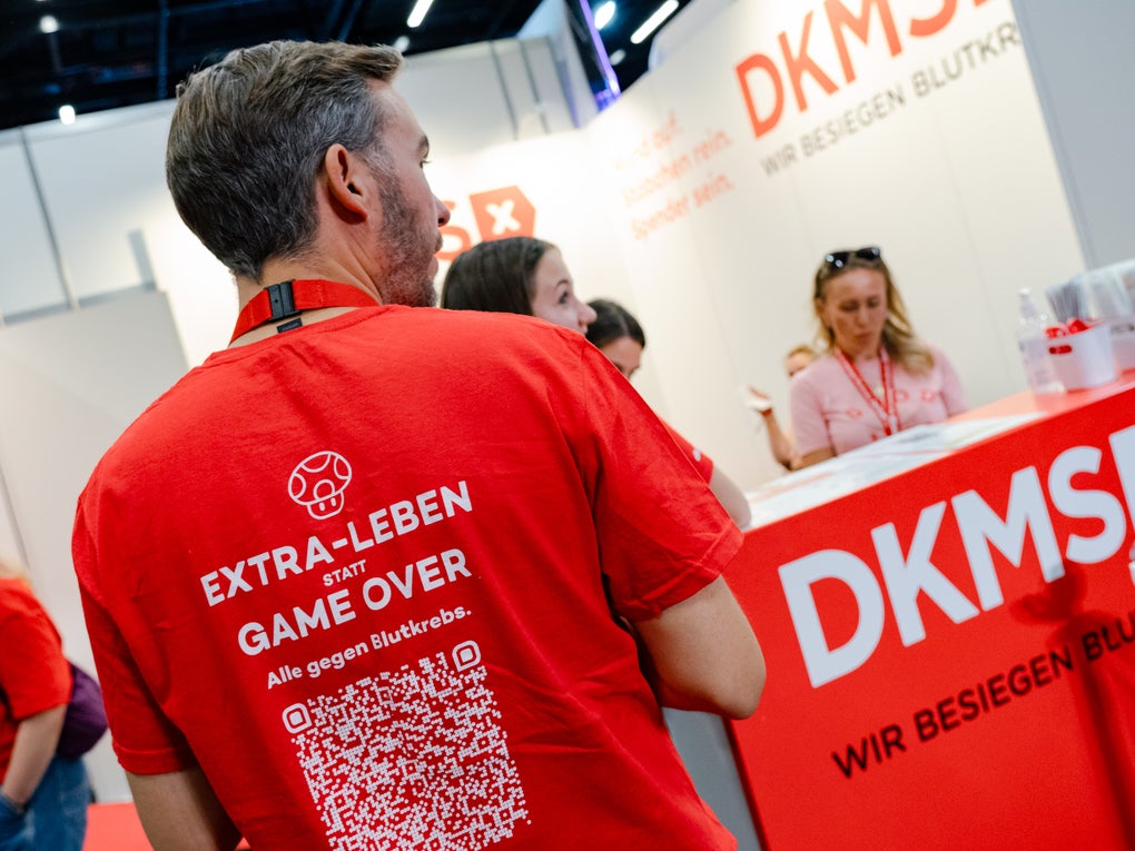 Ein Mann steht mit dem Rücken zur Kamera an einem DKMS-Messestand. Auf seinem roten T-Shirt steht: „EXTRA-LEBEN STATT GAME OVER – Alle gegen Blutkrebs“ sowie ein großer QR-Code. Im Hintergrund sind weitere Personen am DKMS-Stand zu sehen, der mit Informationsmaterial und dem Slogan „Wir besiegen Blutkrebs“ gestaltet ist.
Ein Mann steht mit dem Rücken zur Kamera an einem DKMS-Messestand. Auf seinem roten T-Shirt steht: „EXTRA-LEBEN STATT GAME OVER – Alle gegen Blutkrebs“ sowie ein großer QR-Code. Im Hintergrund sind weitere Personen am DKMS-Stand zu sehen, der mit Informationsmaterial und dem Slogan „Wir besiegen Blutkrebs“ gestaltet ist.
