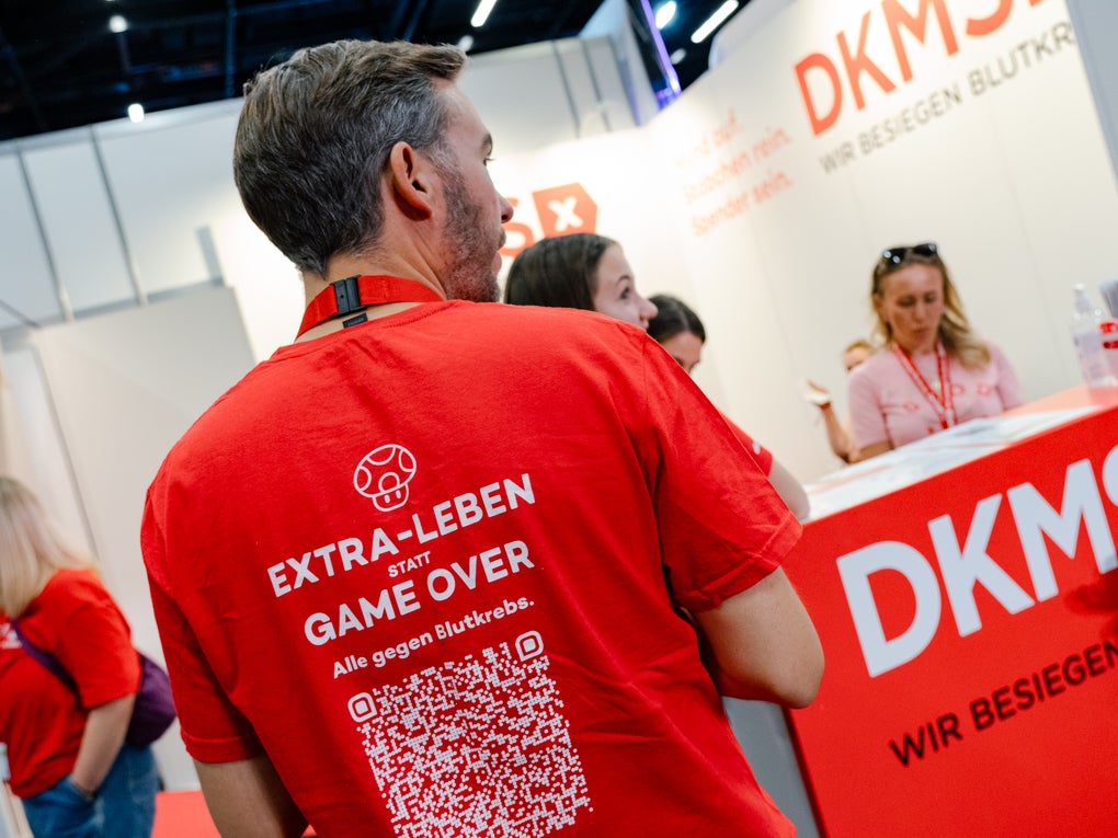 Ein Mann steht mit dem Rücken zur Kamera an einem DKMS-Messestand. Auf seinem roten T-Shirt steht: „EXTRA-LEBEN STATT GAME OVER – Alle gegen Blutkrebs“ sowie ein großer QR-Code. Im Hintergrund sind weitere Personen am DKMS-Stand zu sehen, der mit Informationsmaterial und dem Slogan „Wir besiegen Blutkrebs“ gestaltet ist.
Ein Mann steht mit dem Rücken zur Kamera an einem DKMS-Messestand. Auf seinem roten T-Shirt steht: „EXTRA-LEBEN STATT GAME OVER – Alle gegen Blutkrebs“ sowie ein großer QR-Code. Im Hintergrund sind weitere Personen am DKMS-Stand zu sehen, der mit Informationsmaterial und dem Slogan „Wir besiegen Blutkrebs“ gestaltet ist.
