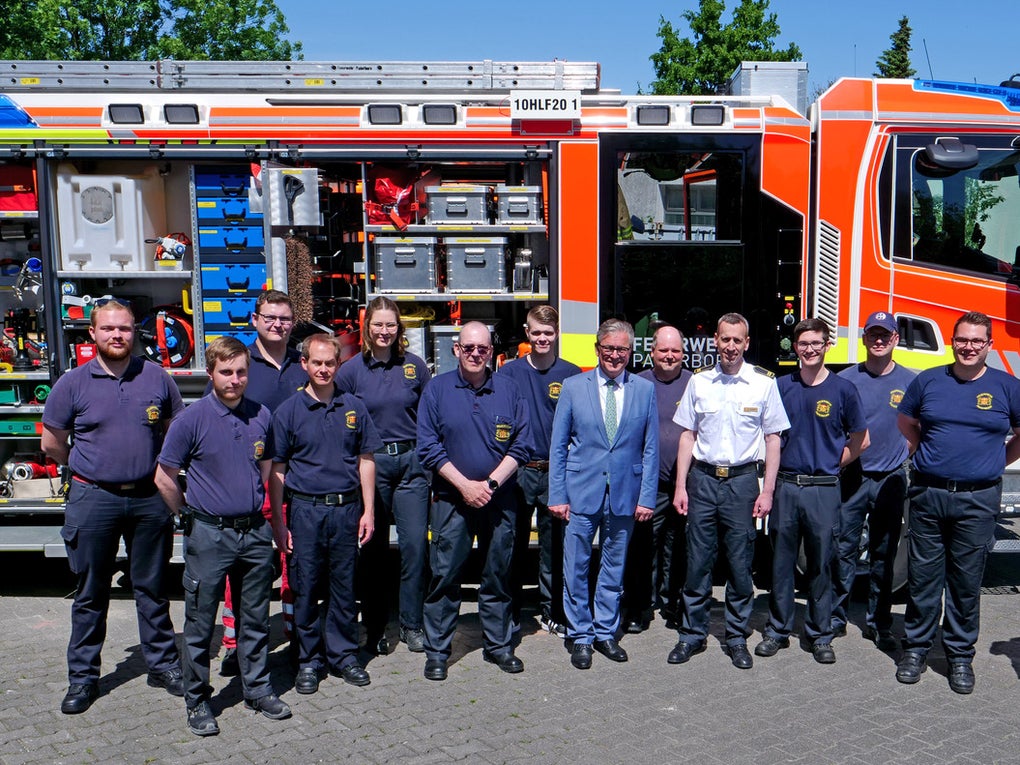 Eine Gruppe von fünfzehn Personen steht in zwei Reihen vor einem Einsatzfahrzeug der Feuerwehr. Die meisten tragen dunkelblaue Einsatzkleidung, einige Personen sind in weißer Dienstkleidung oder einem Anzug gekleidet. Das Einsatzfahrzeug ist orange-weiß mit geöffneten Seitenfächern, in denen Ausrüstung sichtbar ist. Der Hintergrund zeigt ein Gebäude und grüne Bäume bei sonnigem Wetter. Eine Gruppe von fünfzehn Personen steht in zwei Reihen vor einem Einsatzfahrzeug der Feuerwehr. Die meisten tragen dunkelblaue Einsatzkleidung, einige Personen sind in weißer Dienstkleidung oder einem Anzug gekleidet. Das Einsatzfahrzeug ist orange-weiß mit geöffneten Seitenfächern, in denen Ausrüstung sichtbar ist. Der Hintergrund zeigt ein Gebäude und grüne Bäume bei sonnigem Wetter.