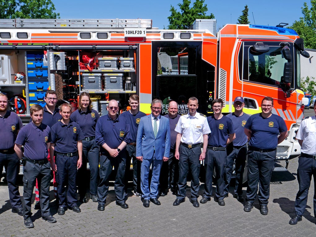 Eine Gruppe von fünfzehn Personen steht in zwei Reihen vor einem Einsatzfahrzeug der Feuerwehr. Die meisten tragen dunkelblaue Einsatzkleidung, einige Personen sind in weißer Dienstkleidung oder einem Anzug gekleidet. Das Einsatzfahrzeug ist orange-weiß mit geöffneten Seitenfächern, in denen Ausrüstung sichtbar ist. Der Hintergrund zeigt ein Gebäude und grüne Bäume bei sonnigem Wetter. Eine Gruppe von fünfzehn Personen steht in zwei Reihen vor einem Einsatzfahrzeug der Feuerwehr. Die meisten tragen dunkelblaue Einsatzkleidung, einige Personen sind in weißer Dienstkleidung oder einem Anzug gekleidet. Das Einsatzfahrzeug ist orange-weiß mit geöffneten Seitenfächern, in denen Ausrüstung sichtbar ist. Der Hintergrund zeigt ein Gebäude und grüne Bäume bei sonnigem Wetter.