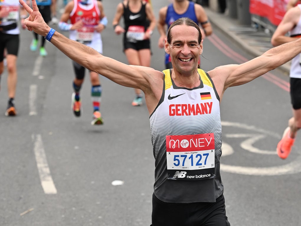 Ein Mann läuft bei einem Straßenmarathon mit ausgestreckten Armen auf die Kamera zu. Er trägt ein Lauftrikot mit der Aufschrift „Germany“, eine Startnummer mit der Ziffer 57127 und ein breites Lächeln im Gesicht. Hinter ihm sind weitere Läuferinnen und Läufer auf der abgesperrten Straße zu sehen.
Ein Mann läuft bei einem Straßenmarathon mit ausgestreckten Armen auf die Kamera zu. Er trägt ein Lauftrikot mit der Aufschrift „Germany“, eine Startnummer mit der Ziffer 57127 und ein breites Lächeln im Gesicht. Hinter ihm sind weitere Läuferinnen und Läufer auf der abgesperrten Straße zu sehen.