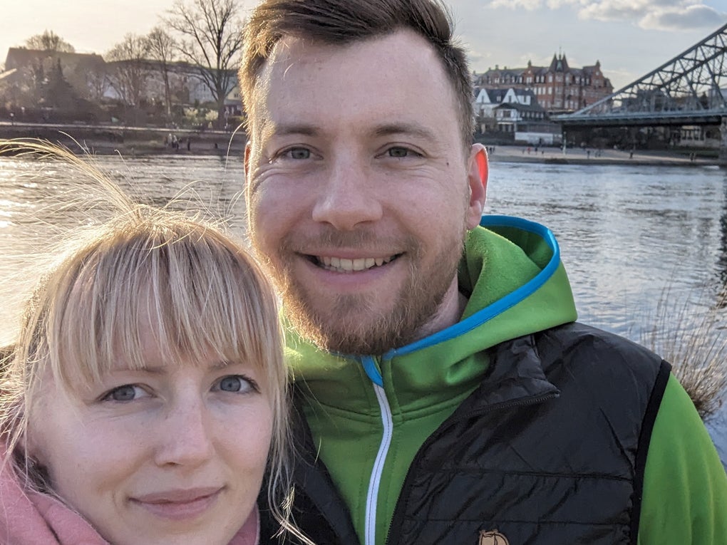Eine Frau und ein Mann lächeln in die Kamera. Sie stehen am Flußufer und im Hintergrund ist eine Brücke zu sehen. Eine Frau und ein Mann lächeln in die Kamera. Sie stehen am Flußufer und im Hintergrund ist eine Brücke zu sehen.