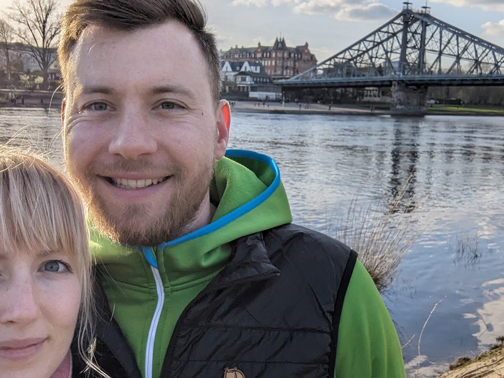 Eine Frau und ein Mann lächeln in die Kamera. Sie stehen am Flußufer und im Hintergrund ist eine Brücke zu sehen. Eine Frau und ein Mann lächeln in die Kamera. Sie stehen am Flußufer und im Hintergrund ist eine Brücke zu sehen.