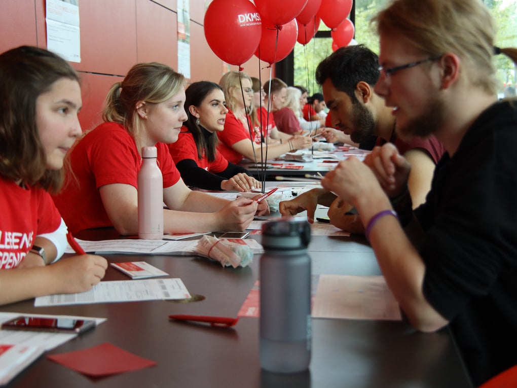 Mehrere junge Menschen in roten DKMS-T-Shirts sitzen an einem langen Tisch und beraten interessierte Personen gegenüber. Auf dem Tisch liegen Infomaterialien und Wasserflaschen. Rote DKMS-Luftballons sind im Hintergrund zu sehen Mehrere junge Menschen in roten DKMS-T-Shirts sitzen an einem langen Tisch und beraten interessierte Personen gegenüber. Auf dem Tisch liegen Infomaterialien und Wasserflaschen. Rote DKMS-Luftballons sind im Hintergrund zu sehen