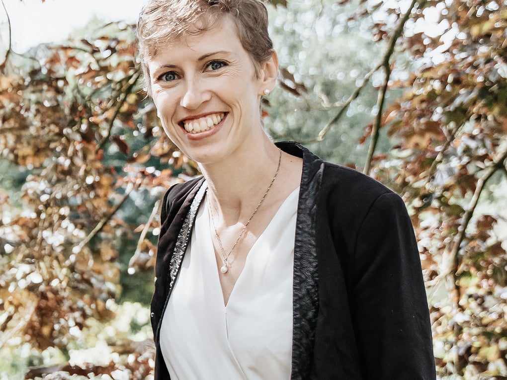 Eine Frau lächelt in die Kamera, im Hintergrund sieht man einen Baum Eine Frau lächelt in die Kamera, im Hintergrund sieht man einen Baum