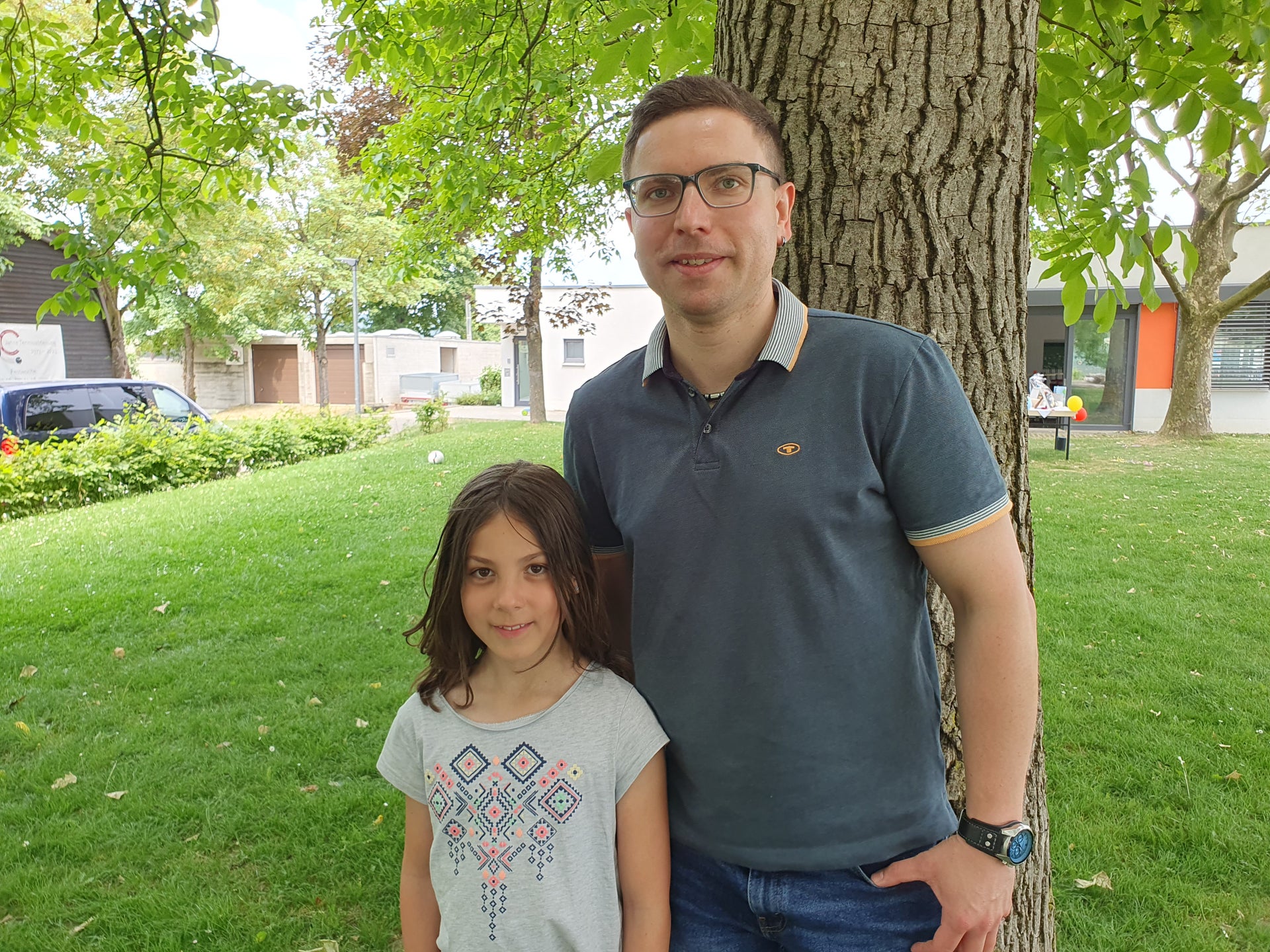 Ein Mann und ein Mädchen stehen nebeneinander auf einer Rasenfläche unter einem Baum. Der Mann trägt ein dunkelgraues Poloshirt und eine Brille, das Mädchen ein helles T-Shirt mit buntem Muster. Im Hintergrund sind Gebäude, ein parkendes Auto und weitere Bäume zu sehen.
Ein Mann und ein Mädchen stehen nebeneinander auf einer Rasenfläche unter einem Baum. Der Mann trägt ein dunkelgraues Poloshirt und eine Brille, das Mädchen ein helles T-Shirt mit buntem Muster. Im Hintergrund sind Gebäude, ein parkendes Auto und weitere Bäume zu sehen.